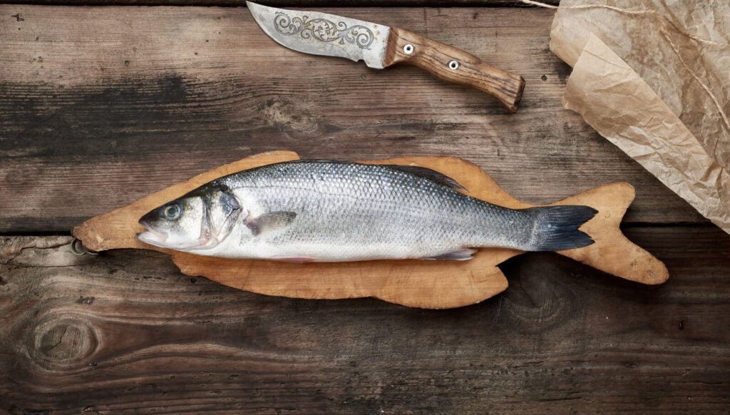 A fish on a wooden cutting board with a knife.